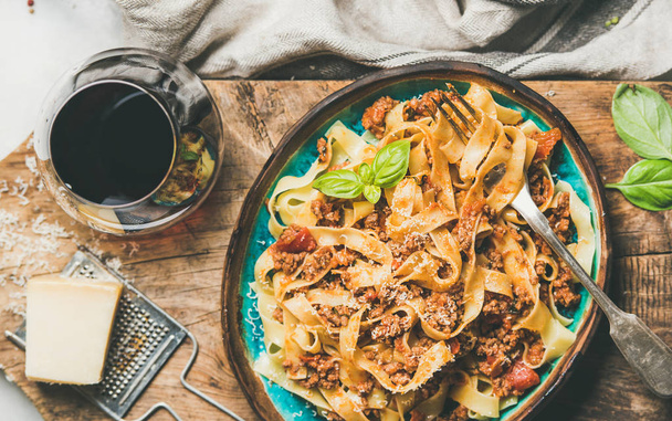 Italian traditional pasta dinner. Flat-lay of Tagliatelle bolognese with minced meat, tomato sauce and grated parmesan cheese and glass of red wine over rustic wooden board background, top view - Foto, Imagem