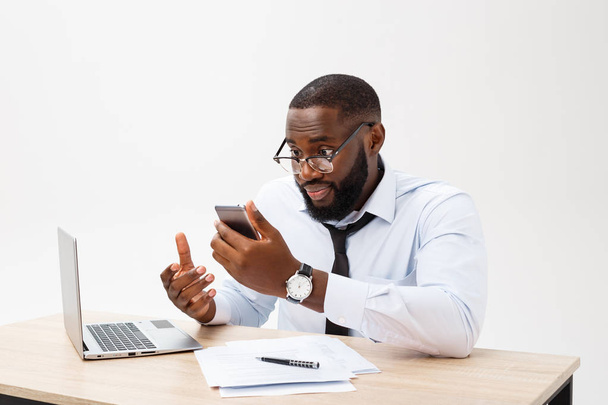 Irritated dark skinned young male enterpreneur being on working place feels very stressed and angry as cannot manage to do all work, surrounded with modern gadgets, looks in displeasure away - 写真・画像