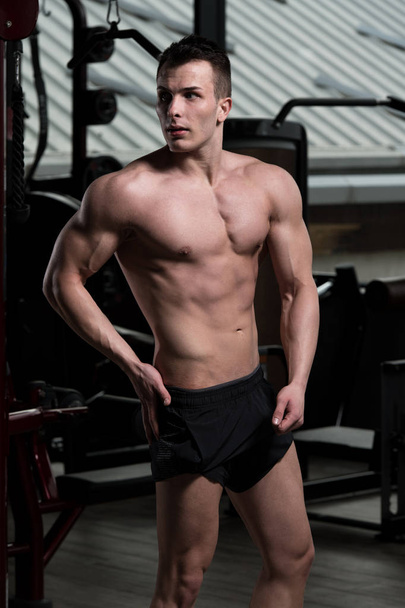 Hombre joven de pie fuerte en el gimnasio y músculos flexionantes - Muscular atlético culturista modelo de fitness posando después de los ejercicios - Foto, imagen