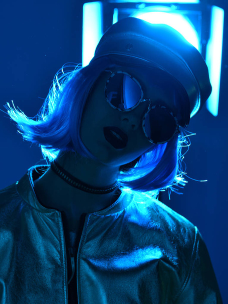 Young beautiful hipster woman in street style fashion winter concept wearing gold hat cap and round sunglasses in blue neon light  - Foto, Imagem