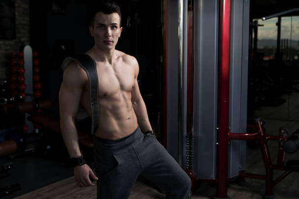 Young Man Standing Strong In The Gym And Flexing Muscles - Muscular Athletic Bodybuilder Fitness Model Posing After Exercises With Belt - Foto, afbeelding
