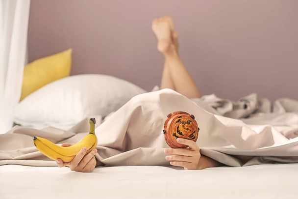 Sleepy woman under blanket with banana and bun in bed - Foto, Imagen