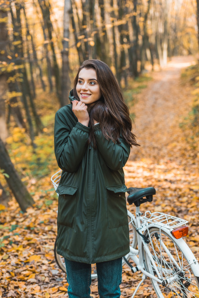 selective focus of smiling young woman looking away near bicycle in autumnal forest - Φωτογραφία, εικόνα