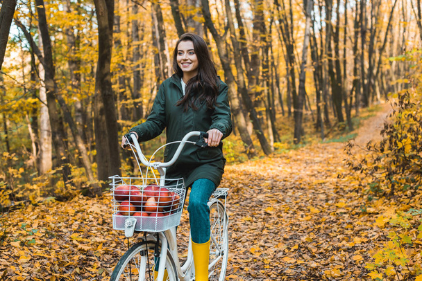 happy girl riding on bicycle with basket full of apples in autumnal forest - Foto, afbeelding