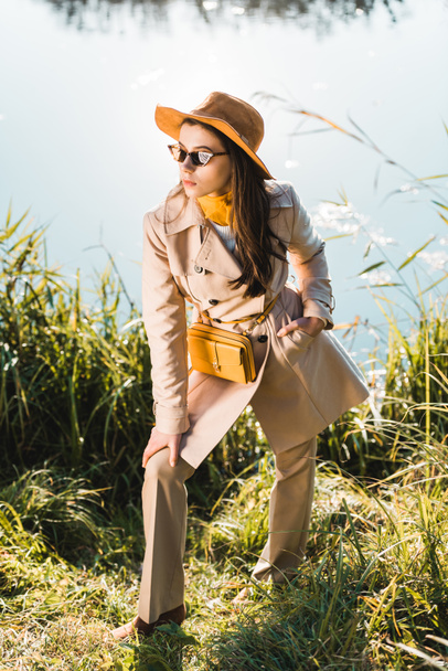 elegant stylish woman in trench coat, hat and sunglasses posing near pond outdoors - Zdjęcie, obraz