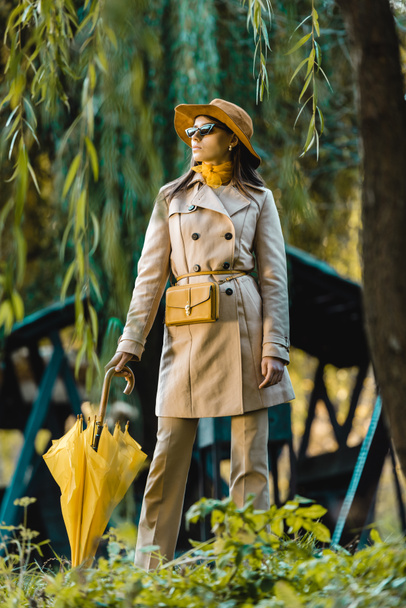 fashionable young woman in trench coat and hat posing with yellow umbrella outdoors - 写真・画像