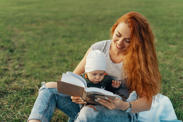 Beautiful mom is reading a book with her cute baby boy and smiling, while sitting on the grass - 写真・画像