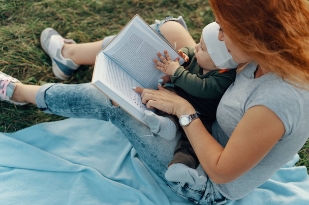 Gyönyörű anya ő aranyos baba fiú egy könyvet olvas, és mosolyogva, miközben ül a fűben. Szög, kilátás. - Fotó, kép