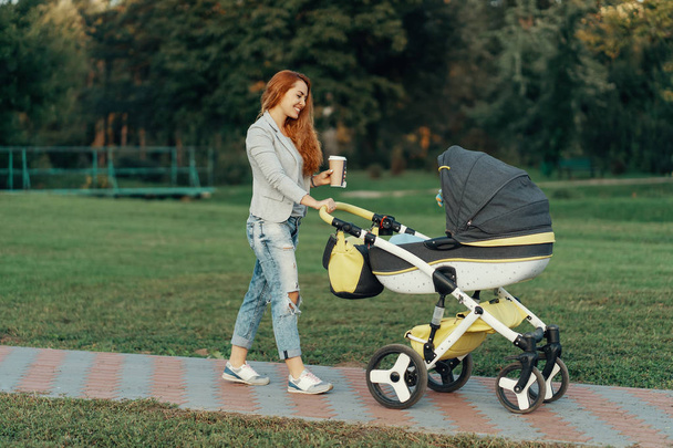 A young mother enjoying the park walk in the morning, drinking a hot tea, with her little child relaxing in his baby trolley. - Фото, зображення