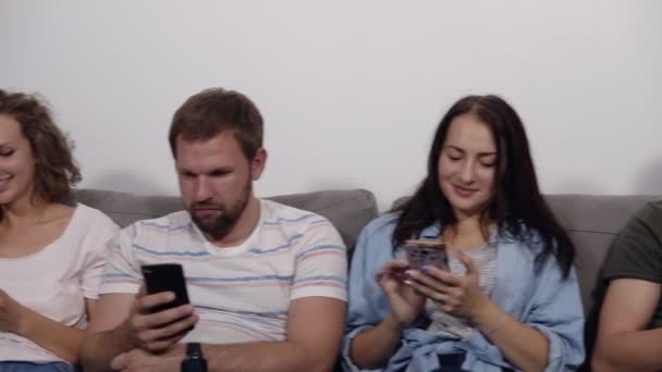 Diverse young smiling people sitting in row on the grey couch together obsessed with devices online, caucasian addicts using their smartphones. White wall background. Front view - Imágenes, Vídeo
