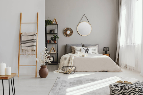 Mirror and clock above bed in bright bedroom interior with pouf and flowers next to ladder. Real photo - Фото, изображение