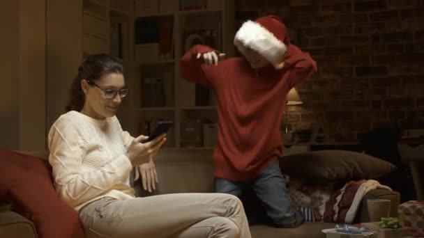 A cheerful boy with Santa hat distracts his mother, she is tickling him and playing, holidays and lifestyle concept - Felvétel, videó