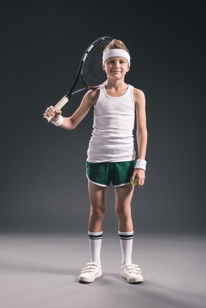 мальчик в спортивной одежде с теннисной ракеткой, смотрящий в камеру на темном фоне
 - Фото, изображение