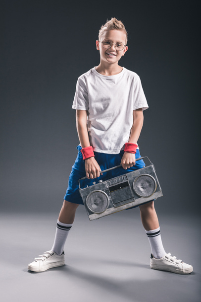 Stylový usměvavý chlapec v brýle s boombox pózuje na šedém pozadí - Fotografie, Obrázek