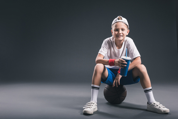 мальчик-подросток с газировкой сидит на баскетбольном мяче на сером фоне
 - Фото, изображение