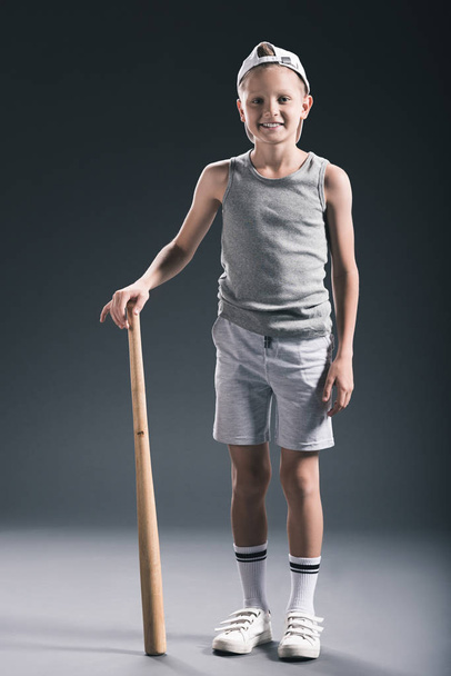 glimlachend jongetje in sportkleding met honkbalknuppel op grijze achtergrond - Foto, afbeelding