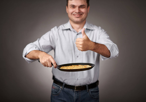 ο άνθρωπος που ποζάρει με μια τηγανίτα στο τηγάνι, άσπρο πουκάμισο και παντελόνι, γκρι φόντο, ρηχό βάθος πεδίου, απότομη τηγανίτα και θαμπή όψη - Φωτογραφία, εικόνα