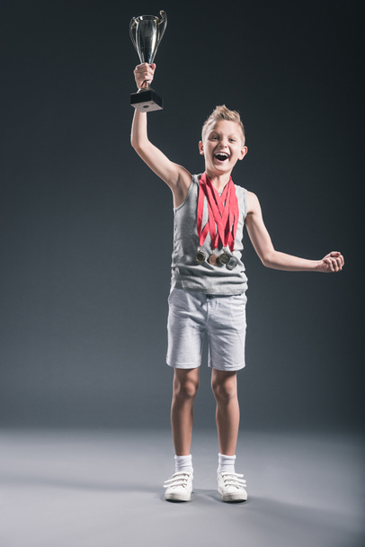 happy boy in sportswear with medals and champions cup gesturing on dark background - Фото, изображение