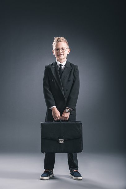 stylish boy in businessman suit and eyeglasses with suitcase on dark background - Foto, Imagem
