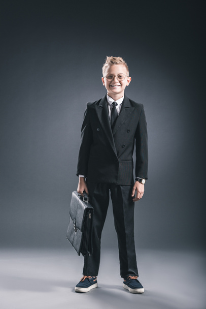 smiling boy in businessman suit and eyeglasses with suitcase on dark background - Fotoğraf, Görsel