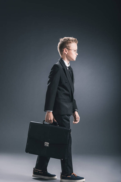 vista lateral de niño preadolescente en traje de hombre de negocios y gafas con maleta sobre fondo oscuro
 - Foto, Imagen
