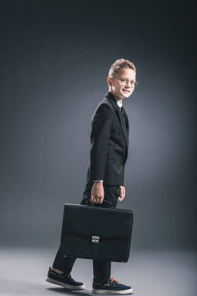vista lateral de niño preadolescente en traje de hombre de negocios y gafas con maleta sobre fondo oscuro
 - Foto, Imagen