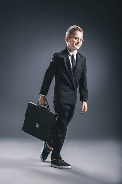 preteen boy in businessman suit and eyeglasses with suitcase walking on dark background - Foto, immagini