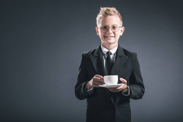 portrait of smiling boy in businessman suit and eyeglasses holding cup of coffee on dark backdrop - Photo, Image