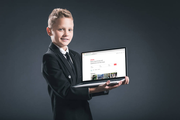 smiling boy in businessman suit showing laptop with airbnb website on screen on grey background - Φωτογραφία, εικόνα