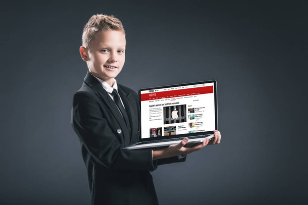 smiling boy in businessman suit showing laptop with bbc website on grey background - Foto, afbeelding