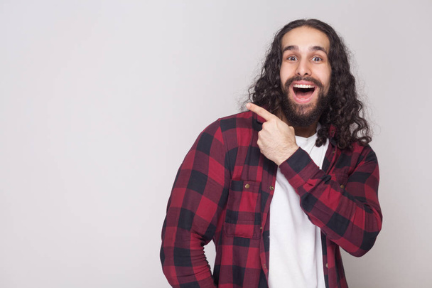 Happy amazed handsome man with beard and black long curly hair in casual style, checkered red shirt looking at camera and pointing at grey background. - 写真・画像