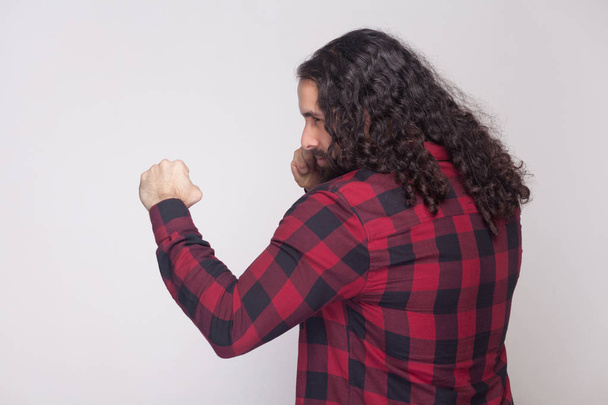 side view portrait of handsome serious man with beard and black long curly hair in checkered red shirt standing with boxing gesture and attacking on grey background. - Foto, immagini