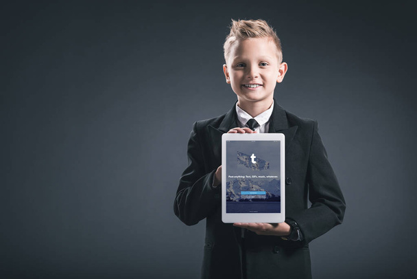 Portrait of smiling boy dressed like businessman showing tablet with tumblr logo in hands on grey backdrop - Photo, Image