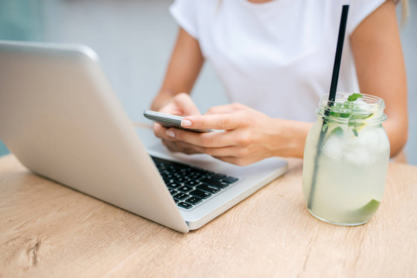 Woman sitting at table with fresh lemonade, laptop and mobile phone. Close-up. - Photo, image