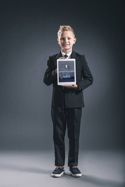 Smiling boy dressed like businessman showing tablet with tumblr logo in hands on grey backdrop - Foto, afbeelding