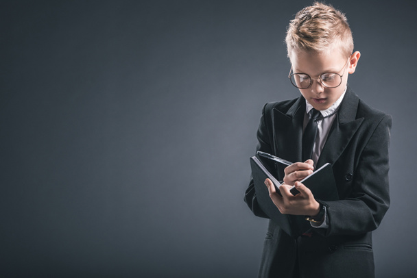 portrait of boy in businessman suit and eyeglasses making notes in notebook on grey backdrop - Фото, зображення