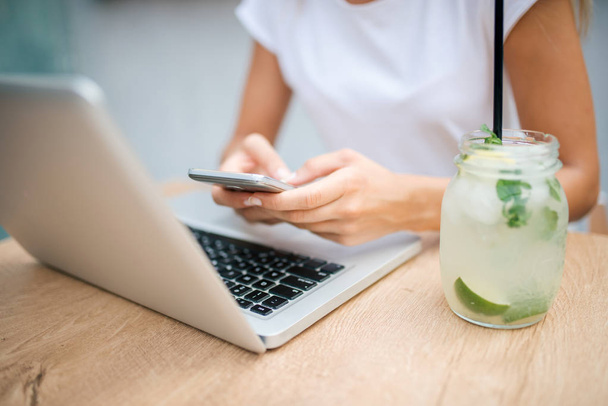 Hands of young woman sitting at table with mobile phone, laptop and fresh lemonade. - 写真・画像