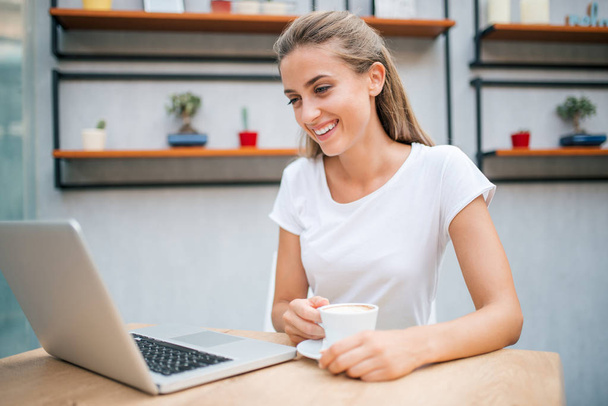Smiling girl looking at laptop while drinking cappuccino. - Φωτογραφία, εικόνα