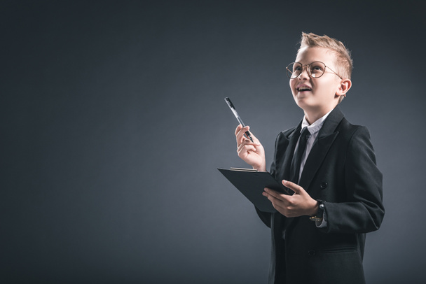 pensive preteen boy in businessman suit and eyeglasses with notepad on grey background - Valokuva, kuva