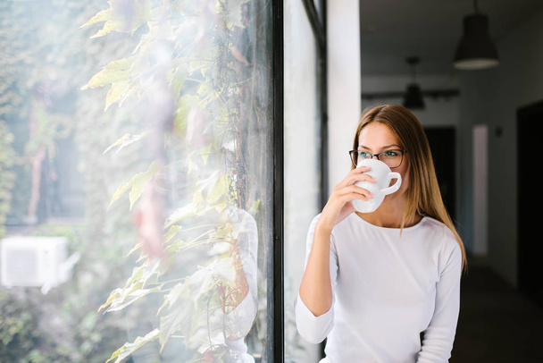 Young woman drinking from a mug near the window.  - Фото, изображение