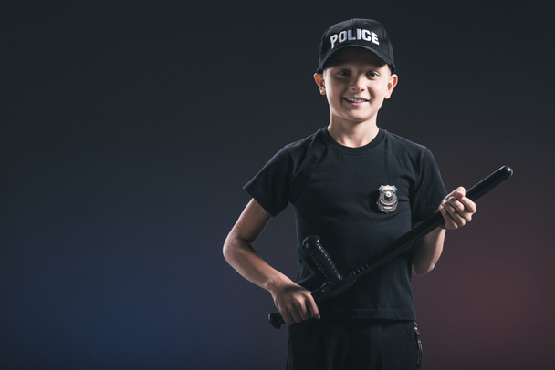smiling preteen boy in policeman uniform with truncheon on dark backdrop - Foto, Imagen
