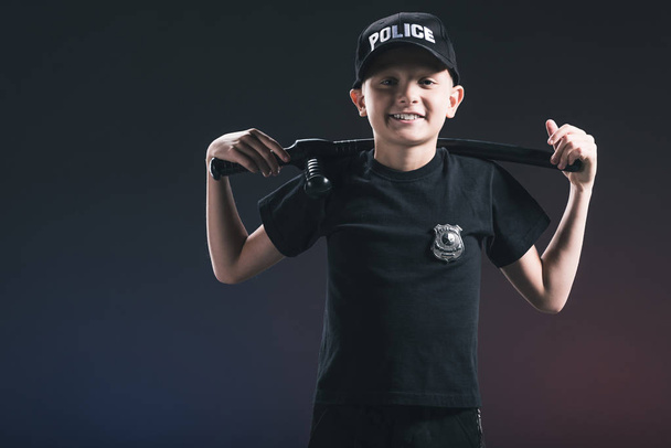 Portret van lachende preteen jongen in politieagent uniform met politieknuppels op donkere achtergrond - Foto, afbeelding