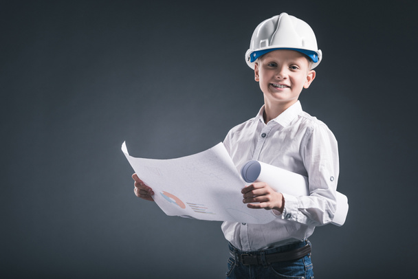 Portret van lachende jongen in architect helm blauwdrukken houden op donkere achtergrond - Foto, afbeelding