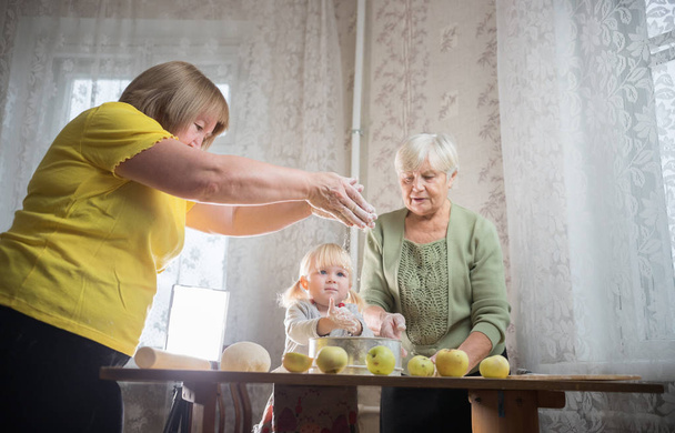 Две пожилые женщины делают яблочные пирожки дома. Просеивание муки с маленькой девочкой
. - Фото, изображение