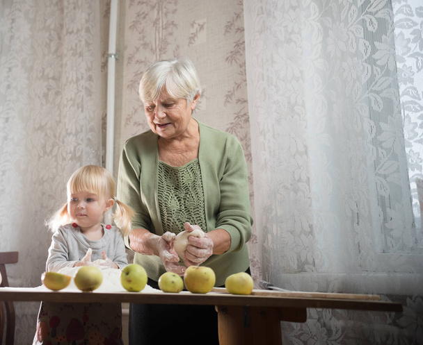 Літня жінка робить яблучні маленькі пироги вдома. Просіяти борошно з маленькою дівчинкою
. - Фото, зображення