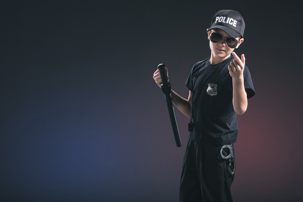 portrait of boy in policeman uniform and sunglasses gesturing on dark background - Фото, изображение