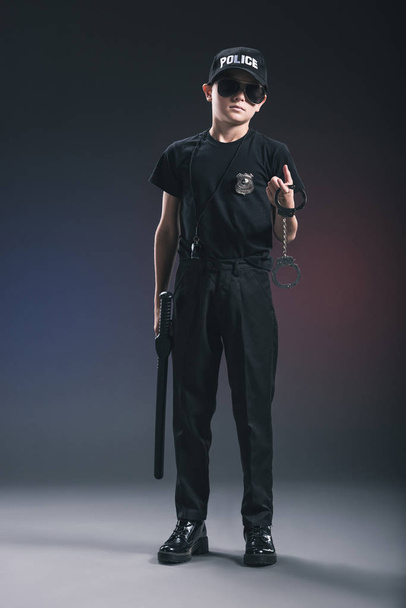 boy in policeman uniform and sunglasses with handcuffs on dark background - Zdjęcie, obraz