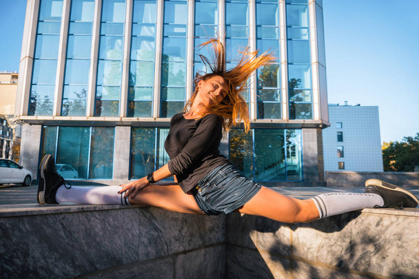 Outdoor lifestyle image of a young pretty chestnut haired caucasian girl doing split, shaking hair and having fun. Cute casual outfit in front of glass building. - Photo, Image