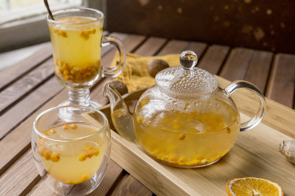 Sea buck thorn tea in glass teapot with glass cups and spoon, orange and walnuts at orange napkin near, at wooden tray on wooden background - Foto, imagen