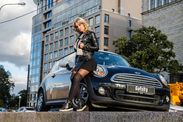 Outdoor portrait of beauty blond woman leaning on modern car. Stylish female model in trendy clothes standing close to little black car  - Photo, Image